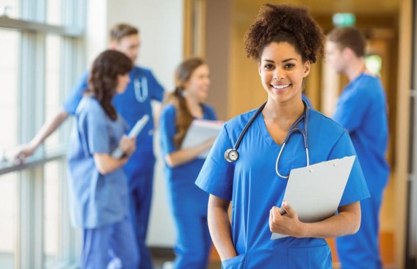 Woman in scrubs holding a clipboard with 4 people in scrubs in the background.