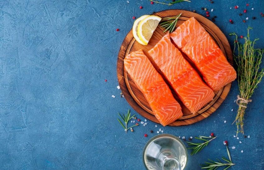 Raw salmon filets on a round wooden board next to herbs and a bottle of oil against a blue surface