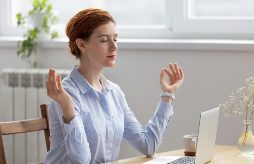 Woman meditating while sitting at a desk in front of a laptop.