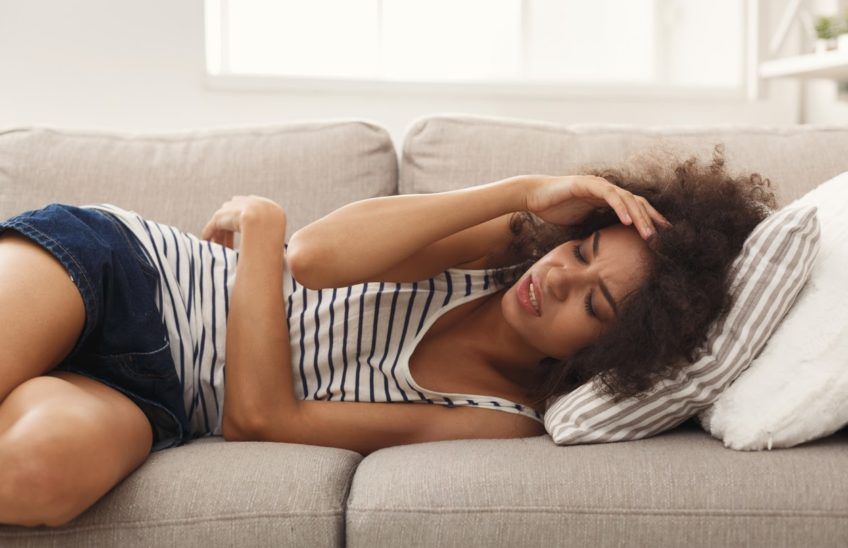 Woman lying on a couch holding her stomach and head in pain.