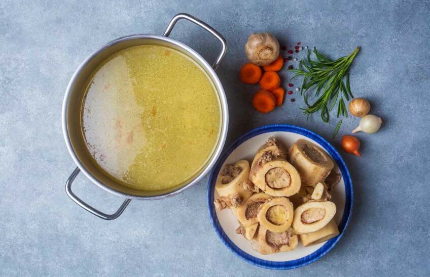 A pot of broth next to cooked bones and fresh vegetables